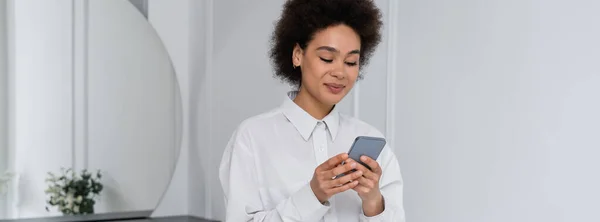 Sonriente mujer afroamericana mensajería en el teléfono inteligente en casa, bandera - foto de stock