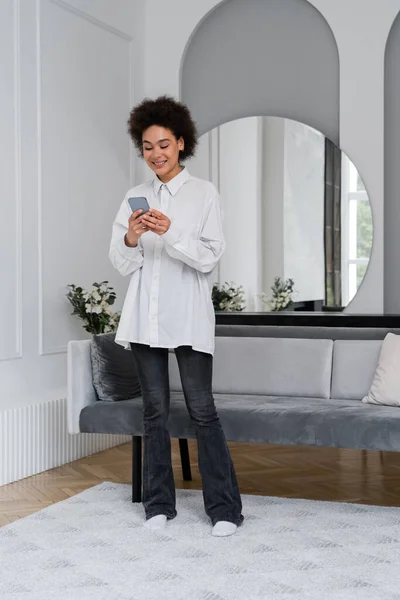 Pleine longueur de messages heureux femme afro-américaine sur smartphone dans le salon moderne — Photo de stock