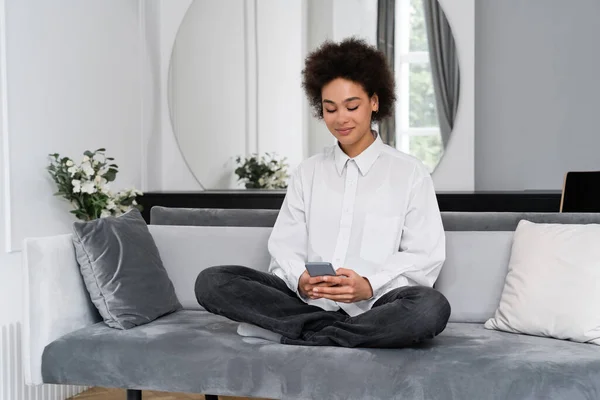 Sorrindo Africano americano mulher mensagens no smartphone enquanto sentado no sofá de veludo — Fotografia de Stock