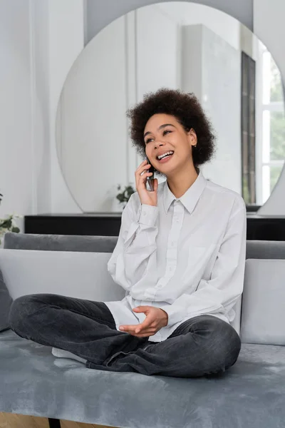 Mujer afroamericana feliz hablando en el teléfono inteligente mientras está sentado en el sofá de terciopelo - foto de stock