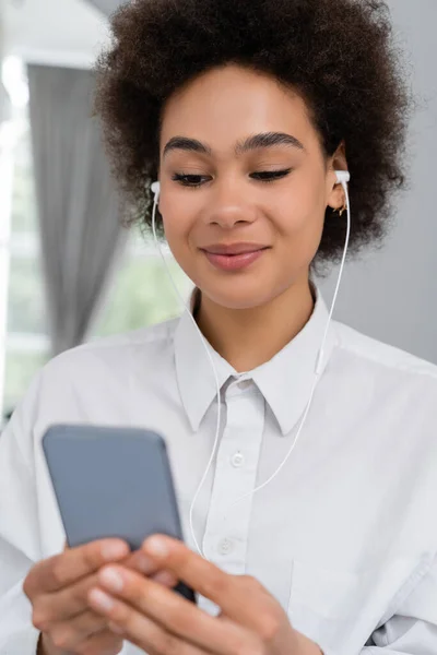 Donna afroamericana sorridente che ascolta musica in cuffia cablata e utilizza smartphone — Foto stock