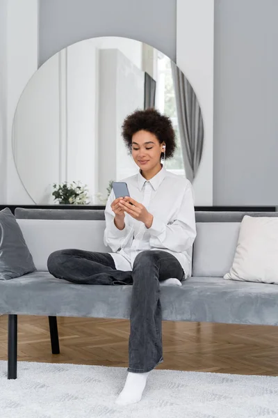 Pleased african american woman listening music in wired earphones and using smartphone in modern living room — Stock Photo