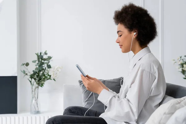 Vista laterale di piacere donna afro-americana ascoltare musica e utilizzando smartphone in soggiorno — Foto stock