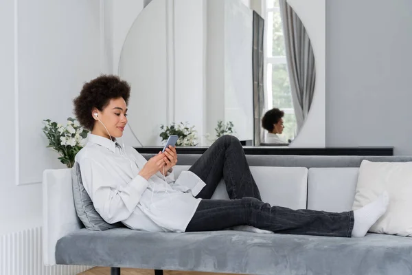 Alegre mujer afroamericana escuchando música en auriculares con cable y el uso de teléfonos inteligentes en la sala de estar moderna - foto de stock