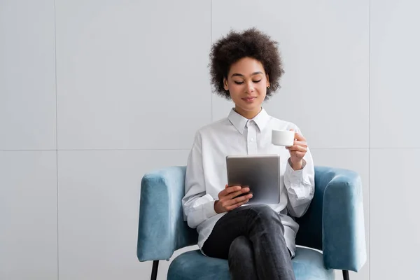 Mujer afroamericana feliz sosteniendo la taza de café y viendo la película en la tableta digital - foto de stock