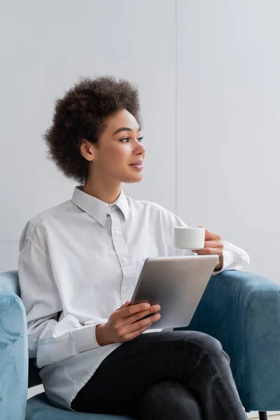 Heureuse femme afro-américaine tenant tasse de café et tablette numérique tout en étant assis dans un fauteuil en velours bleu — Photo de stock