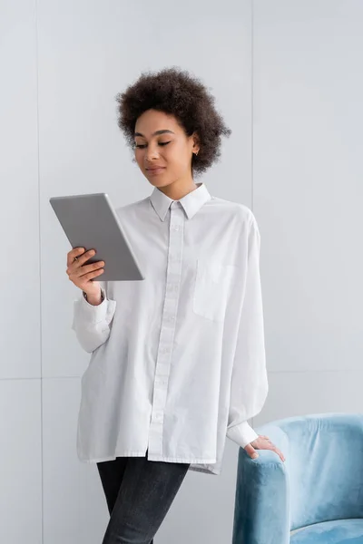 Sorrindo mulher afro-americana segurando tablet digital enquanto estava perto de poltrona de veludo azul — Fotografia de Stock