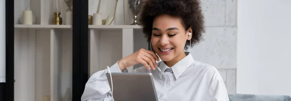 Mujer afroamericana feliz en auriculares cableados sosteniendo la tableta digital mientras mira la película, bandera - foto de stock