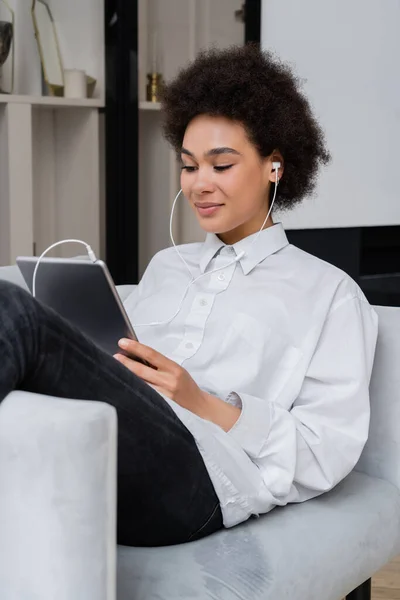 African american woman in wired earphones holding digital tablet while watching movie and sitting on grey armchair — Stock Photo