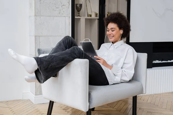 Glückliche afrikanisch-amerikanische Frau mit Kopfhörern, die ein digitales Tablet hält, während sie sich einen Film ansieht und auf einem grauen Sessel sitzt — Stockfoto