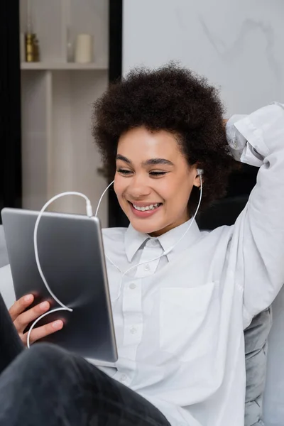 Alegre afroamericana mujer en auriculares con cable celebración de la tableta digital mientras mira la película de comedia - foto de stock