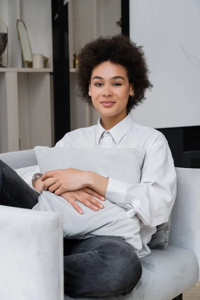 Donna afroamericana sorridente e riccia in camicia bianca con colletto seduta con cuscino su poltrona grigia — Foto stock