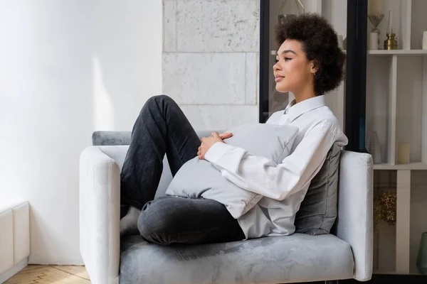 Dreamy and curly african american woman in white shirt with collar sitting with pillow on grey armchair — Stock Photo