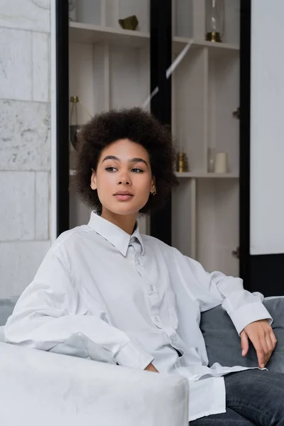 Mujer afroamericana soñadora y rizada en camisa blanca con cuello sentado en sillón de terciopelo gris - foto de stock