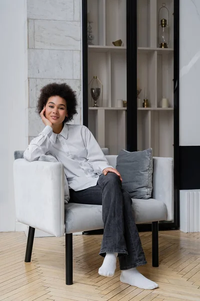 Smiling and curly african american woman in white shirt with collar sitting on grey modern armchair — Stock Photo