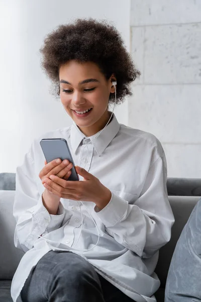 Mujer afroamericana feliz en auriculares con cable usando teléfono móvil - foto de stock