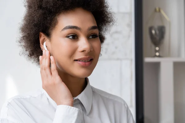 Retrato de mujer afroamericana feliz escuchando música y ajustando auriculares inalámbricos - foto de stock