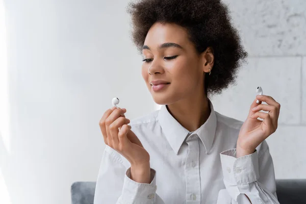 Happy african american woman listening music and looking at wireless earphone — Stock Photo