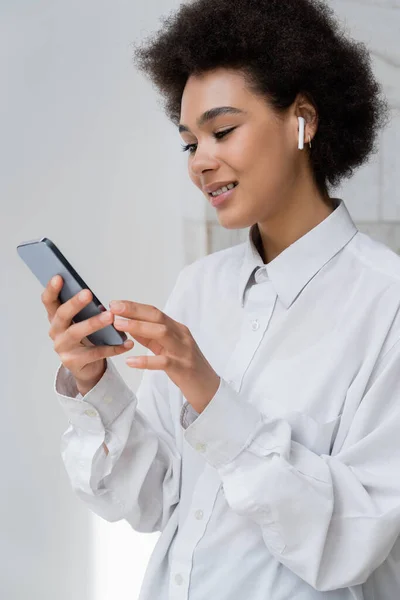 Mulher americana africana feliz ouvindo música no fone de ouvido sem fio e usando smartphone — Fotografia de Stock