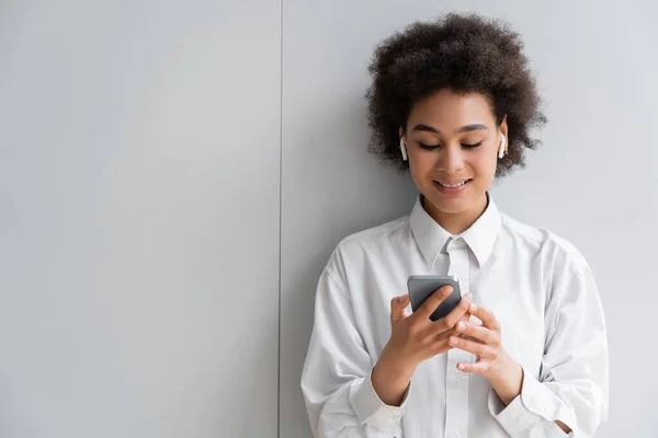 Mulher africana feliz ouvindo música em fones de ouvido sem fio e usando smartphone perto da parede cinza — Fotografia de Stock