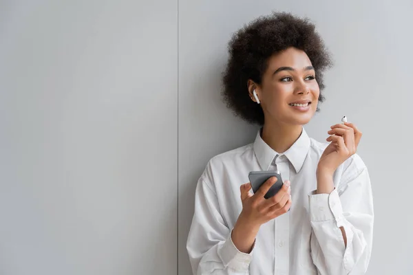 Cheerful african american woman listening music in wireless earphone and holding smartphone near grey wall — Stock Photo