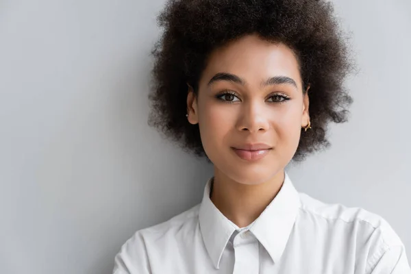 Portrait of curly african american woman in white shirt with collar standing near grey wall — Stock Photo