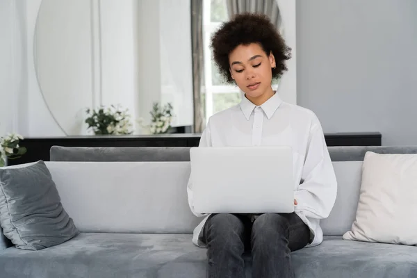 Mujer afroamericana que usa el ordenador portátil mientras trabaja desde casa en la sala de estar moderna - foto de stock