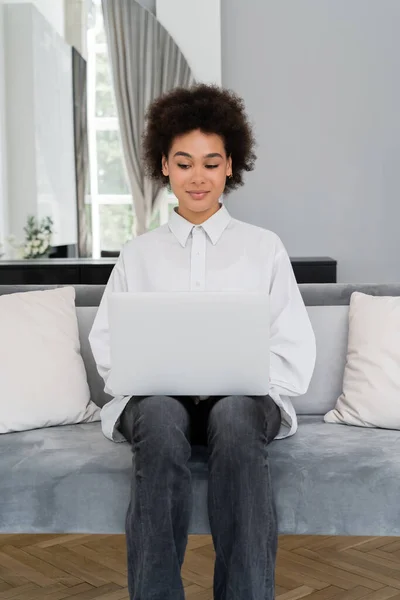 Femme afro-américaine souriante utilisant un ordinateur portable tout en étant assis sur un canapé en velours gris — Photo de stock