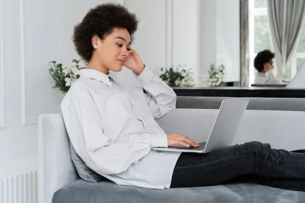 Mulher americana africana usando laptop enquanto sentado no sofá de veludo cinza e trabalhando em casa — Fotografia de Stock