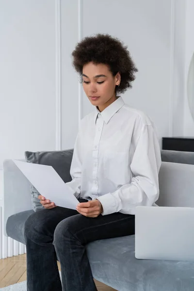 Mulher afro-americana olhando para o documento em branco perto do laptop no sofá cinza veludo — Fotografia de Stock