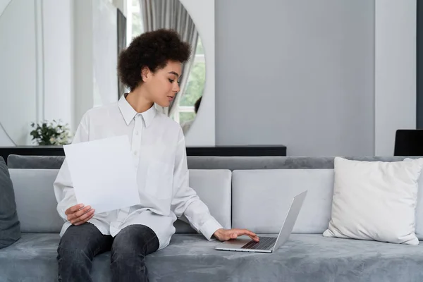 Mujer afroamericana mirando el documento en blanco y usando el ordenador portátil en el sofá gris terciopelo - foto de stock