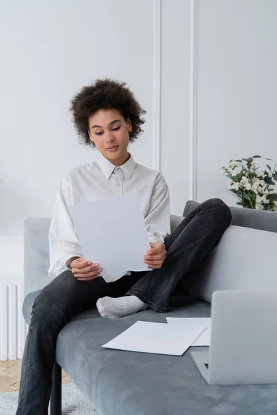 Femme afro-américaine frisée regardant un document vierge près d'un ordinateur portable sur un canapé gris velours — Photo de stock