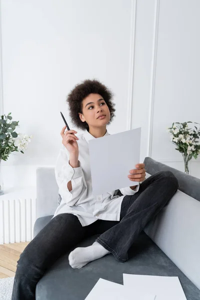 Mujer afroamericana pensativa sosteniendo pluma y documento en blanco mientras está sentada en un sofá gris - foto de stock