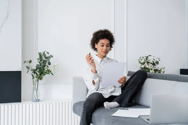 Encaracolado Africano americano mulher segurando caneta e documento em branco enquanto sentado no sofá cinza perto do laptop — Fotografia de Stock