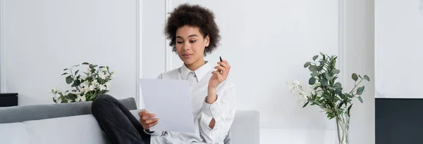 Mujer afroamericana rizada sosteniendo pluma y documento en blanco mientras trabaja desde casa, pancarta - foto de stock