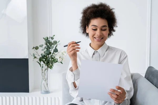 Sorrindo mulher afro-americana segurando caneta e documento em branco enquanto trabalhava em casa — Stock Photo