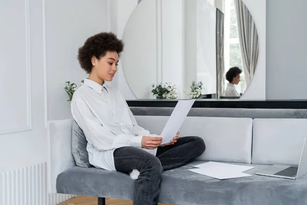 Mujer afroamericana rizada sosteniendo documento en blanco cerca de la computadora portátil en el sofá mientras trabaja desde casa - foto de stock