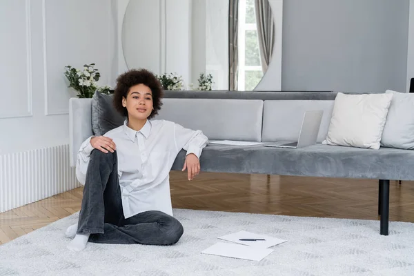 Mujer afroamericana rizada sentada cerca de la computadora portátil y documentos en la alfombra - foto de stock