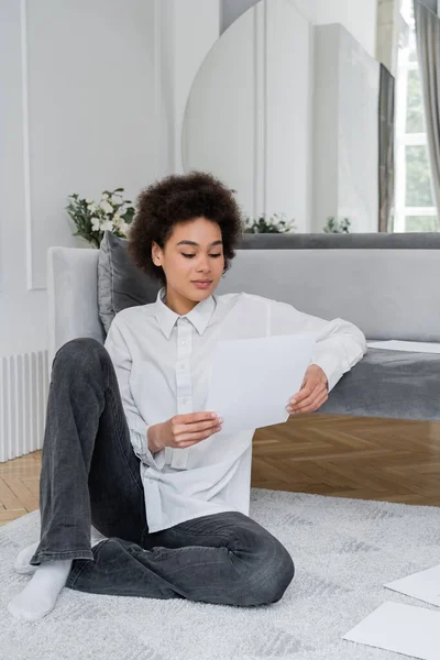 Femme afro-américaine bouclée tenant un document vierge tout en travaillant à la maison et assis près du canapé en velours — Photo de stock