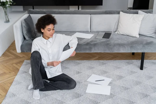 Mujer afroamericana sosteniendo documento en blanco mientras está sentada cerca de la computadora portátil en un sofá de terciopelo - foto de stock