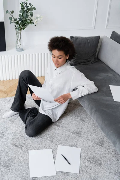 Vue grand angle de la femme afro-américaine tenant un document vierge tout en étant assis sur le tapis près du canapé en velours gris — Photo de stock