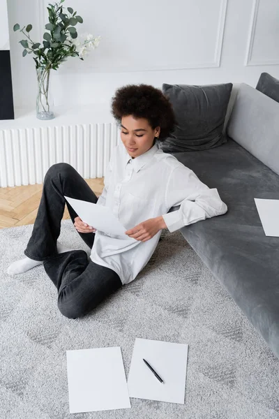 Hochwinkelaufnahme einer afrikanisch-amerikanischen Frau mit leerem Dokument, während sie auf einem Teppich neben einem grauen Samtsofa sitzt — Stockfoto