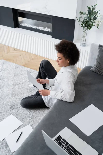 Vue grand angle de la femme afro-américaine tenant le document tout en étant assis sur le tapis près de l'ordinateur portable sur le canapé — Photo de stock