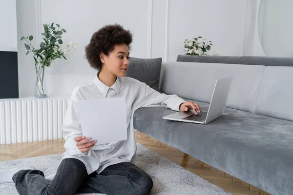 Curly african american freelancer holding paper and using laptop on grey velvet sofa — Stock Photo