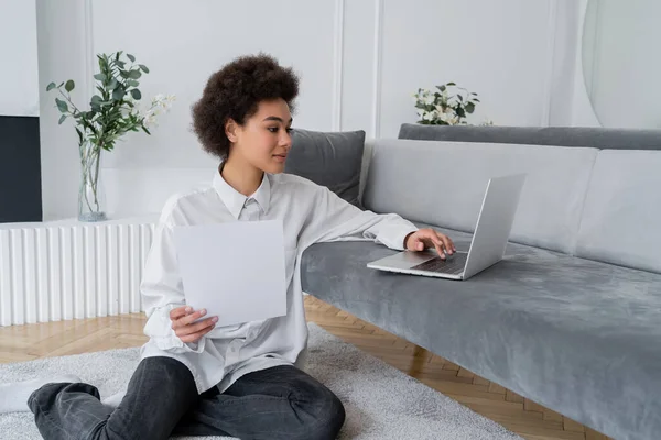 Rizado afroamericano freelancer celebración de papel y el uso de ordenador portátil en sofá de terciopelo gris - foto de stock