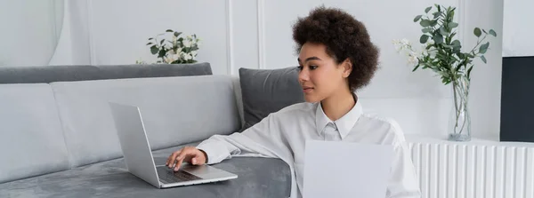 Curly african american freelancer holding paper and using laptop on grey velvet sofa, banner — Stock Photo