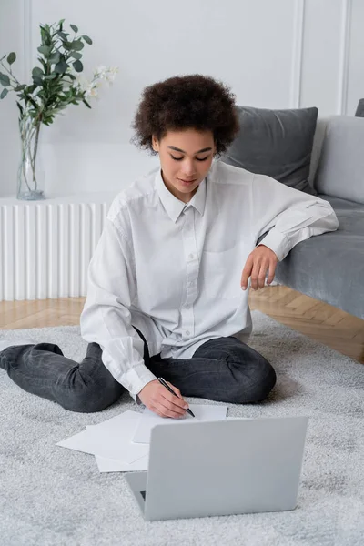 Mujer afroamericana escribiendo en papel mientras está sentada en la alfombra cerca de la computadora portátil - foto de stock