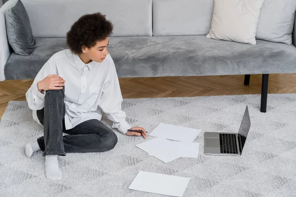 Vista lateral de la mujer afroamericana mirando a la computadora portátil mientras está sentado en la alfombra cerca de documentos - foto de stock