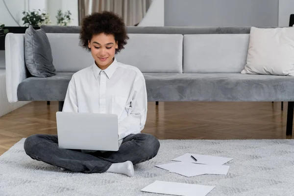 Afrikanerin lächelt, während sie Laptop in der Nähe von Dokumenten auf Teppich benutzt — Stockfoto