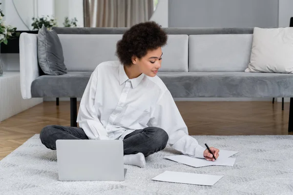 African american woman smiling while writing on paper and sitting with laptop on carpet — Stock Photo